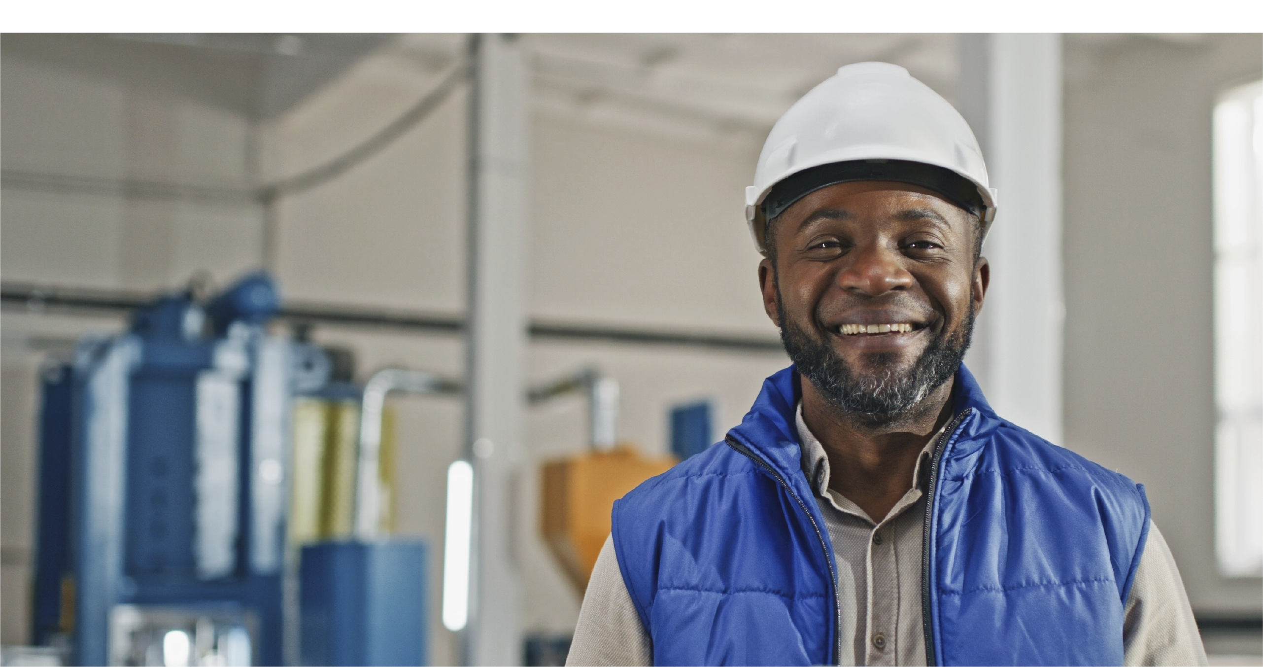 Smiling worker in a hard hat and blue vest standing confidently in an industrial facility, highlighting professionalism and a safe workplace environment